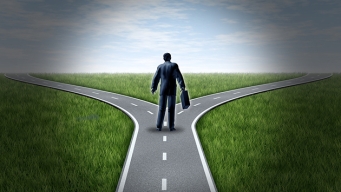 Business man at a cross roads standing at a horizon with grass and blue sky showing a fork in the road representing the concept of a strategic dilemma choosing the right direction to go when facing two equal or similar options.